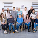 Gruppenfoto von 15 Personen, die in einem hellen Innenraum auf Stühlen sitzen und stehen.